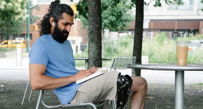 olive skinned man with long dark hair and a prosthetic leg is reading while sitting at a table outside.jpg.optimal