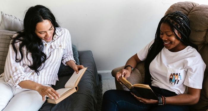 two women one with light tan skin and the other with brown skin reading books and laughing with each other.jpg.optimal