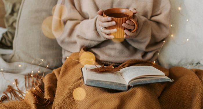 a fair skinned person holding a mug with a book and autumn themed decor in their lap.jpg.optimal