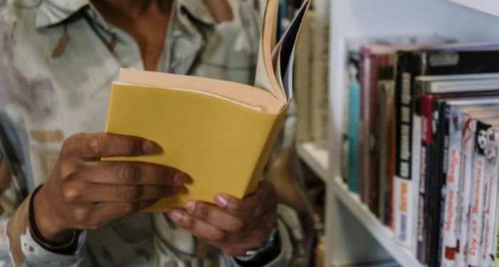 a brown skinned person holding a book with a yellow cover while standing next to a book shelf.jpg.optimal