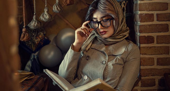 a lighter skinned woman in a head scarf and glasses reading while leaning against a brick wall.jpg.optimal