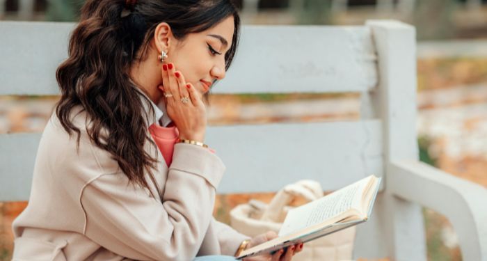 a lightly tan skinned woman sitting pleasantly reading a book while sitting on a bench 1.jpg.optimal